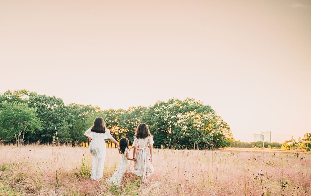Three kids in light summer dresses stand in tall pink‑gold grasses during sunset, facing the distant tree line, creating a peaceful environmental photography scene with warm natural light and soft autumn tones.