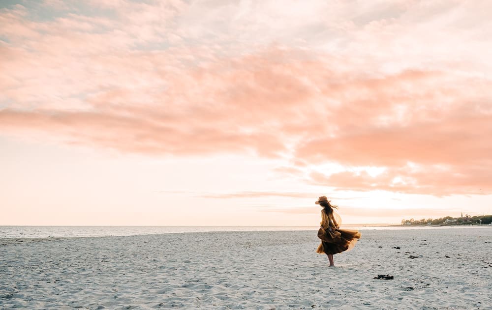 A person in a mustard yellow flowy dress and brown brimmed hat stands slightly bent toward the ocean, alone in a wide shot with soft natural light and an open shoreline, creating a quiet environmental photography moment. Captured in a wide shot.