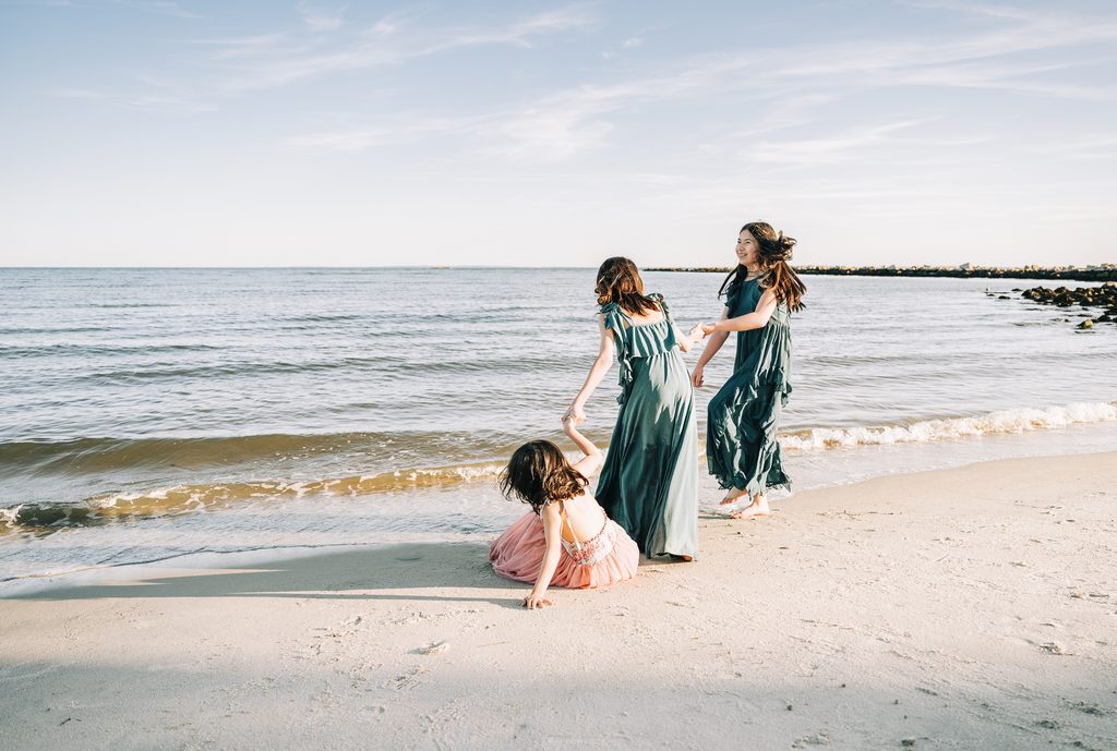 Three people at the shoreline in a candid storytelling photography moment, with two standing in teal dresses helping a seated person in a pink dress by the ocean waves.