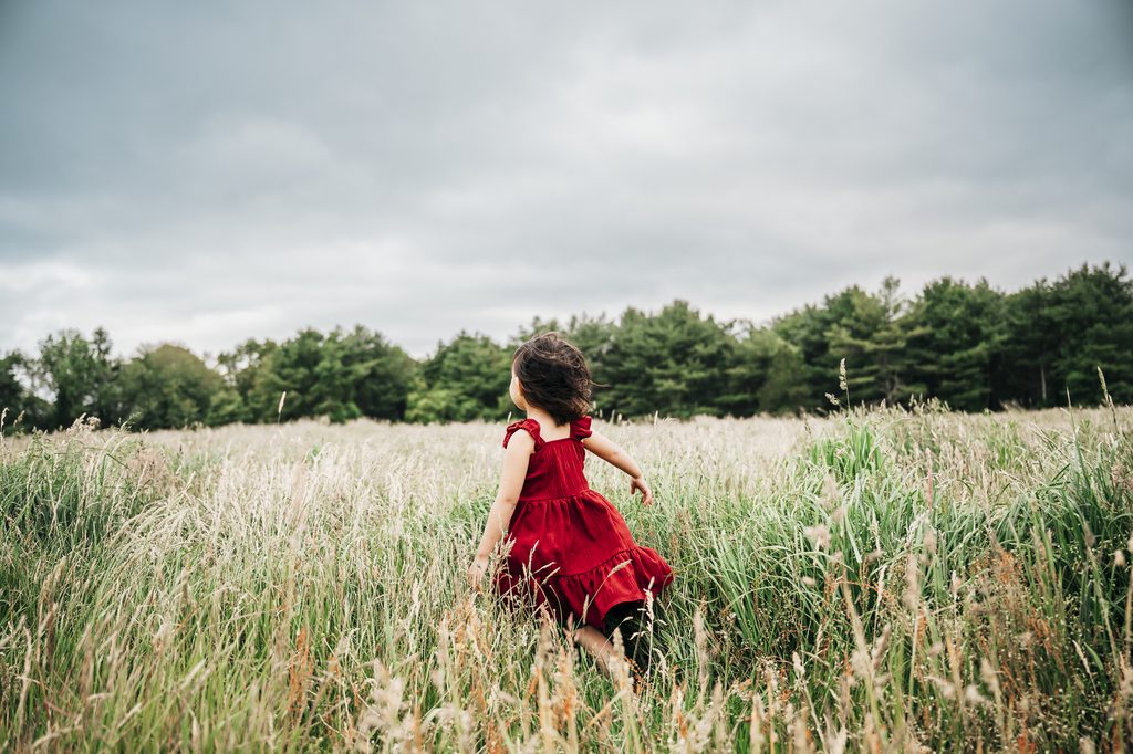 Child in a red dress exploring a field of tall grass in a natural storytelling photography moment with trees and a cloudy sky in the background.