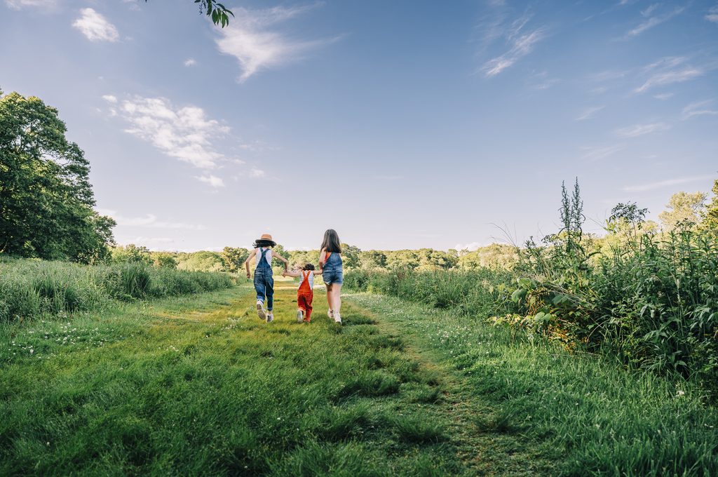 Three people walking along a grassy nature trail in a storytelling photography scene, surrounded by lush greenery and a bright blue sky.